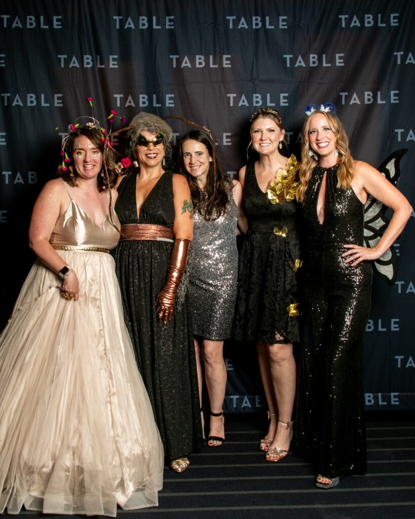 Five women in sparkling attire and whimsical headpieces pose for a photo event.