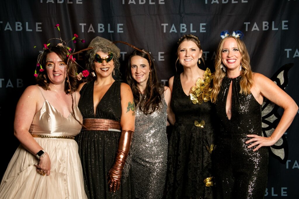 Five women in sparkly and whimsical outfits pose for a photo at an event with "TABLE" branding.