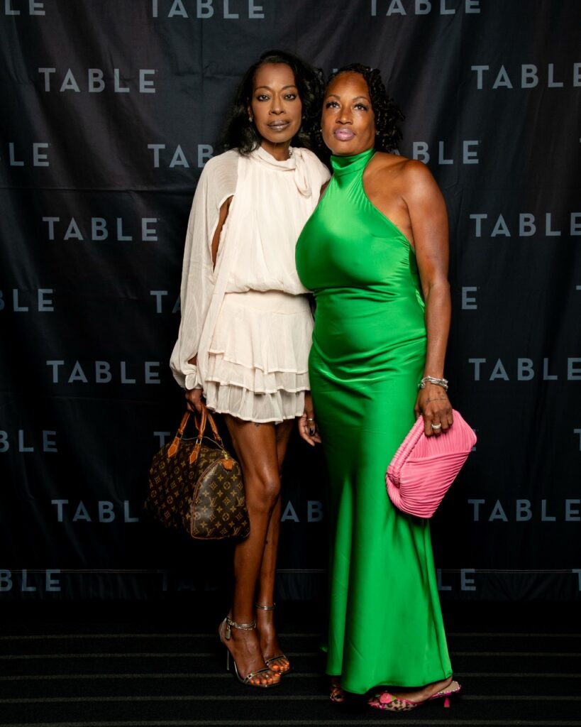 Two women in formal attire pose in front of a black backdrop with "TABLE" repeated.