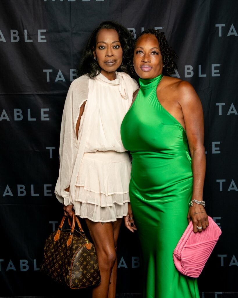 Two stylish women in elegant dresses pose in front of a backdrop with the word "TABLE" repeated.