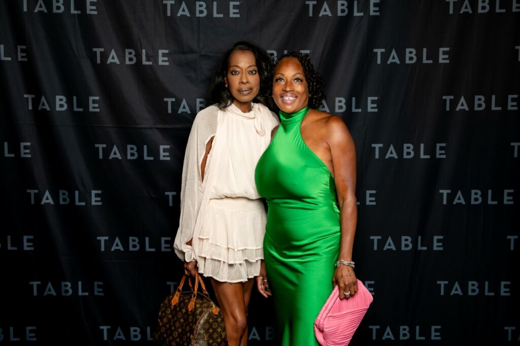 Two women in formal attire pose in front of a black backdrop with "TABLE" repeated.