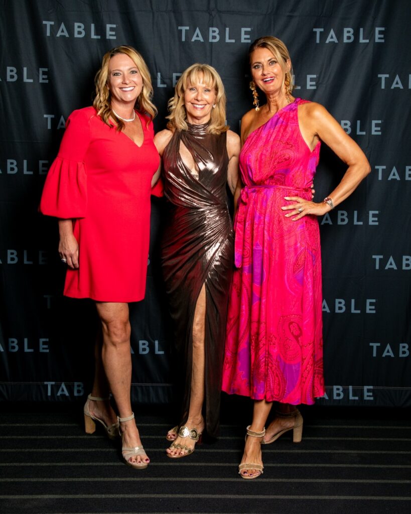Three women in formal dresses pose for a photo at an event with "TABLE" branding.