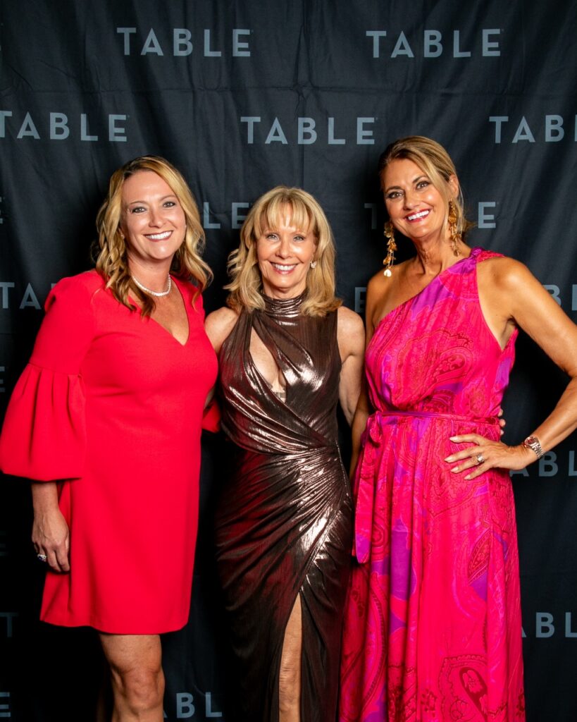 Three women in elegant dresses pose for a photo at an event with a "TABLE" backdrop.