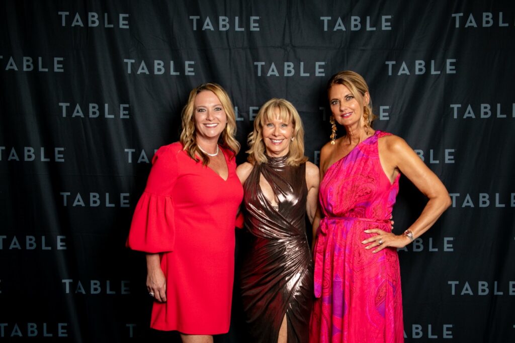 Three women in formal dresses pose for a photo in front of a black backdrop with "TABLE" repeatedly printed on it.