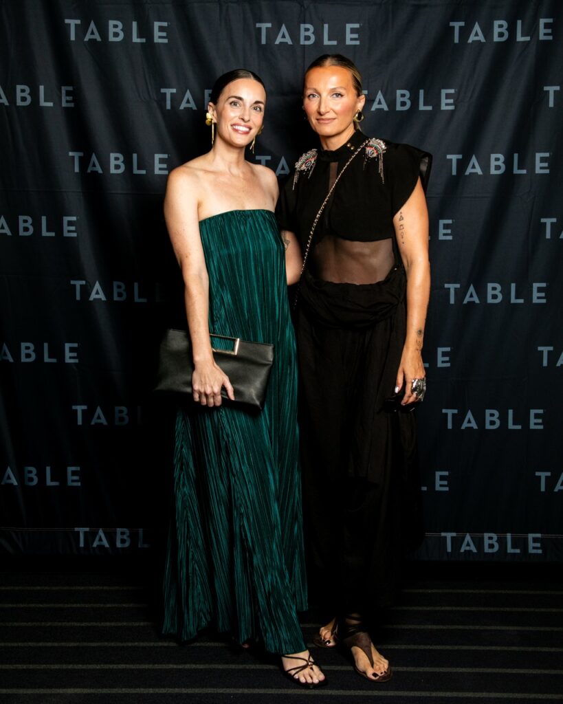 Two stylish women pose in front of a black backdrop with repeating "TABLE" text.