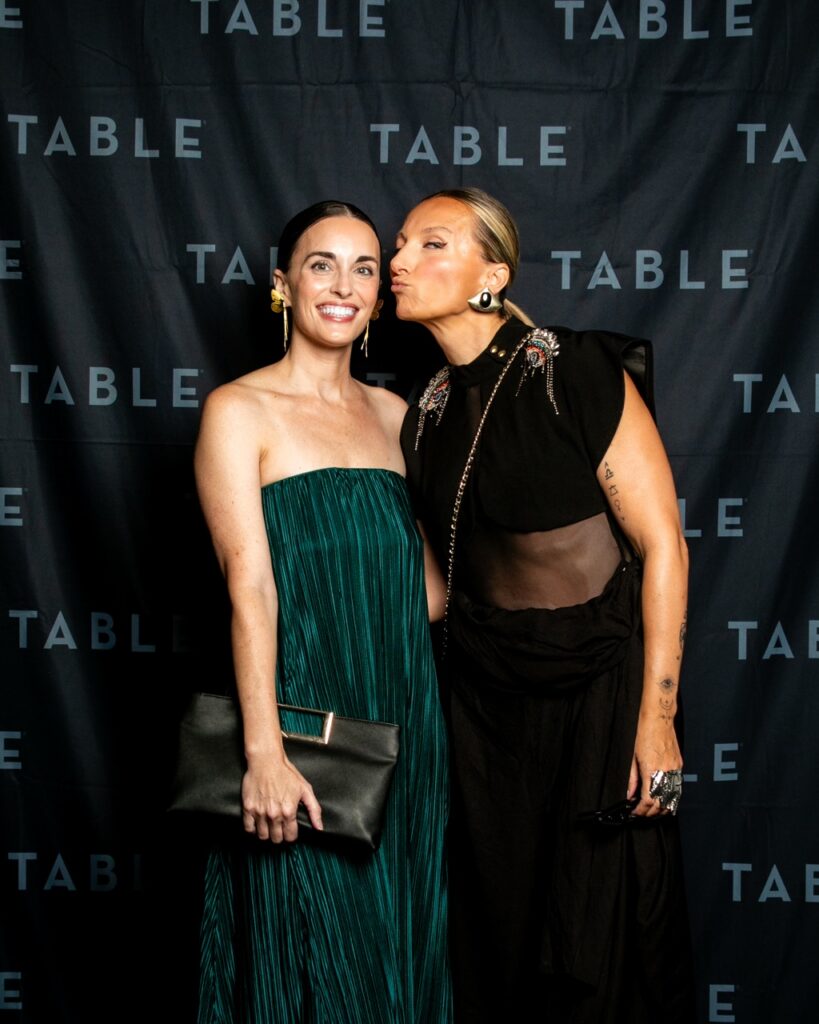 Two women pose for a photo in front of a black backdrop with the word TABLE repeated.