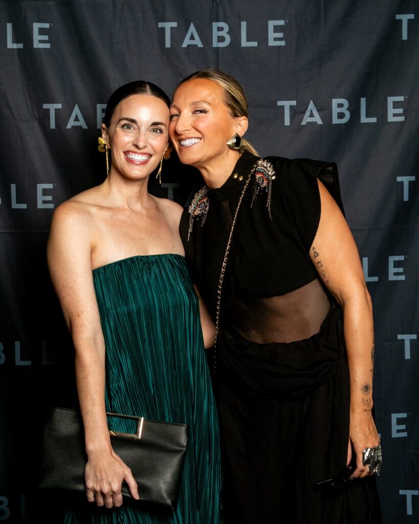 Two women in formal attire smile at the camera in front of a TABLE backdrop.