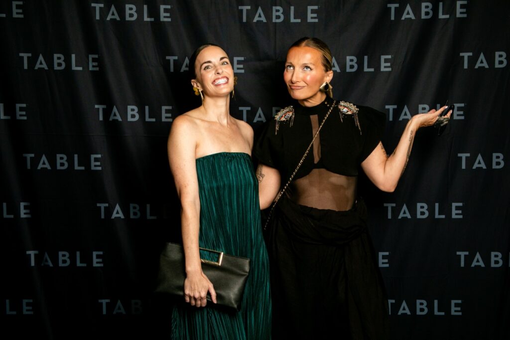 Two women pose for a photo in front of a black backdrop with the word "TABLE" repeated.