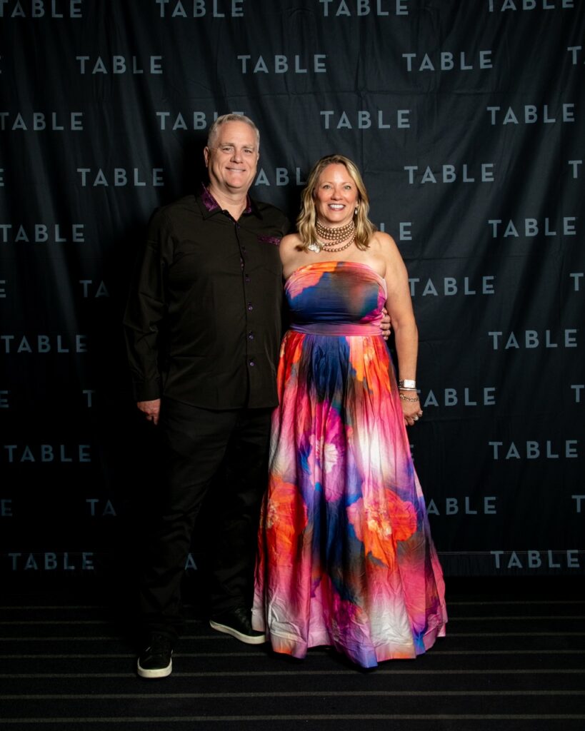 Man and woman in formal attire pose in front of a "TABLE" backdrop.