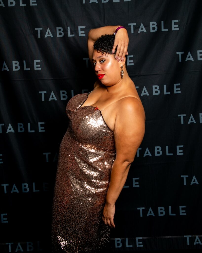 Woman in a sparkling gold dress poses against a black backdrop with "TABLE" repeating