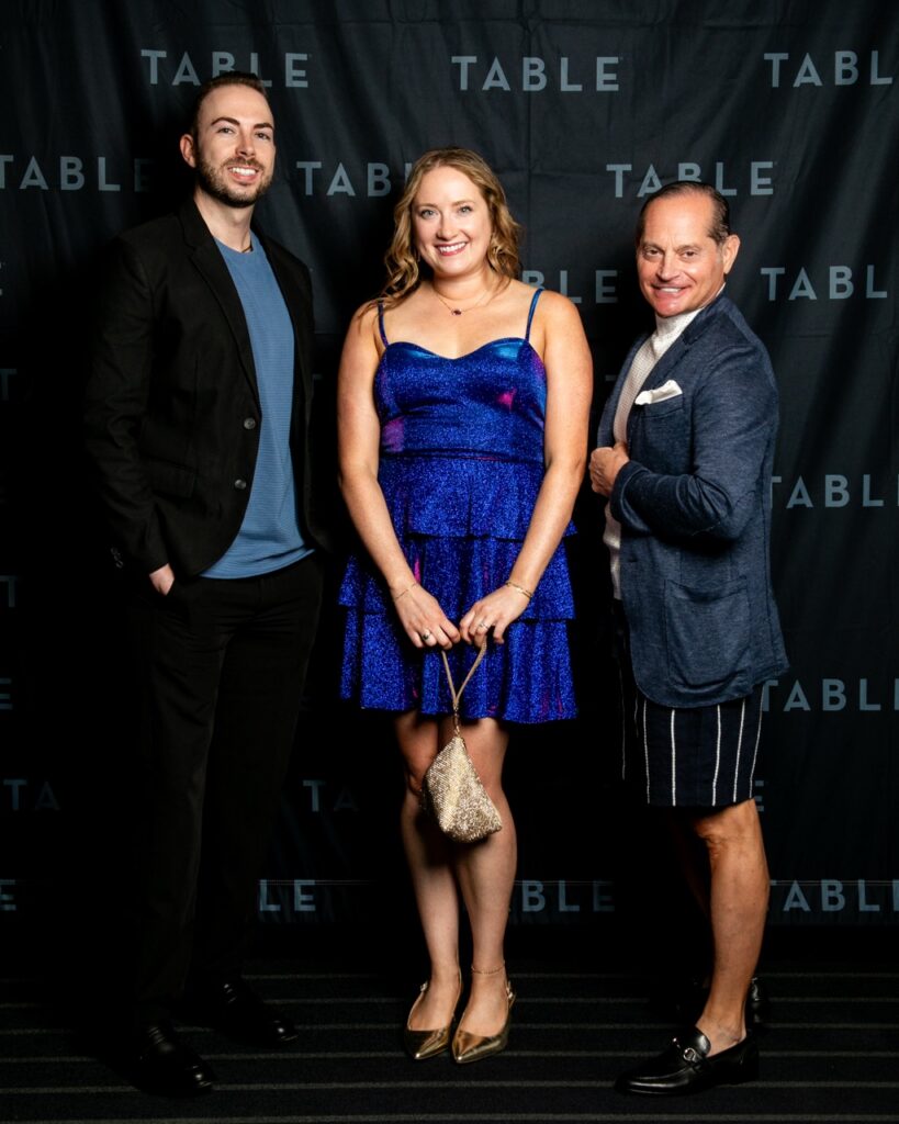 Three people pose for a photo at a TABLE event, two men in suits and a woman in a blue dress.