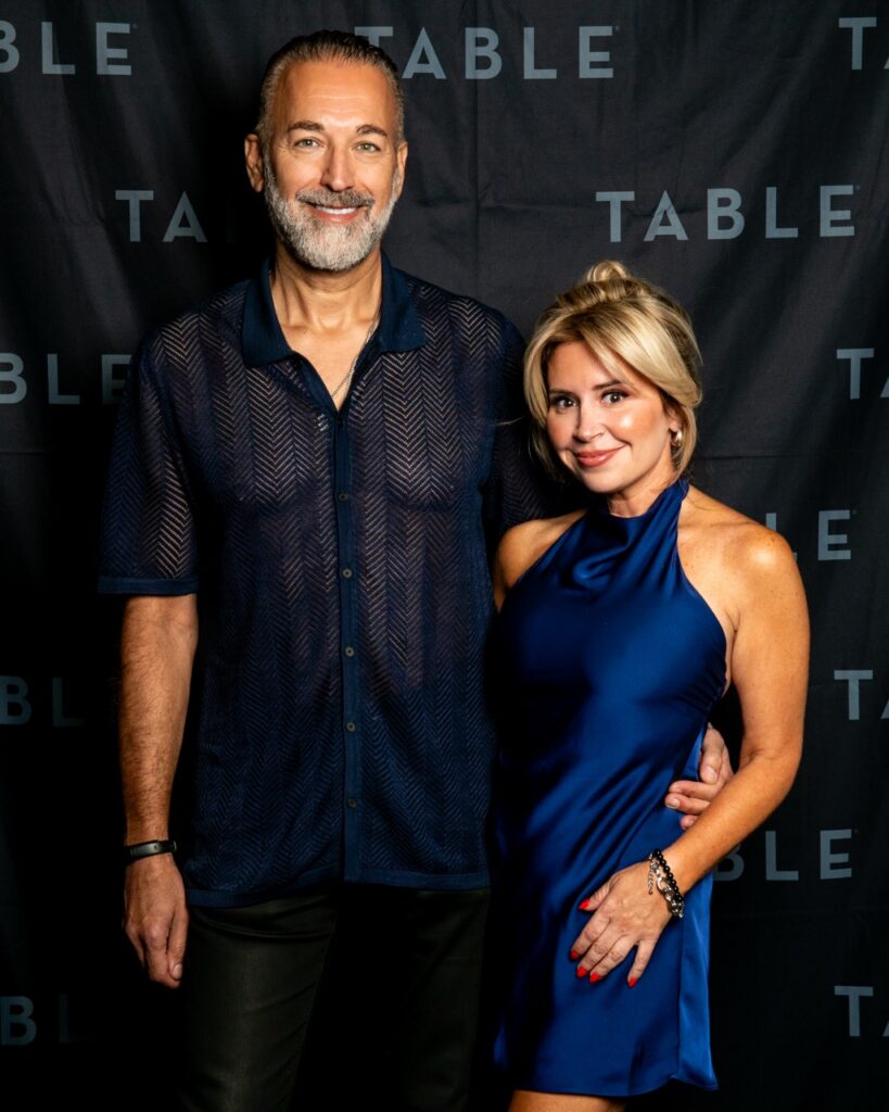 Man and woman posing for a photo in front of a black backdrop with the word "TABLE" repeated.