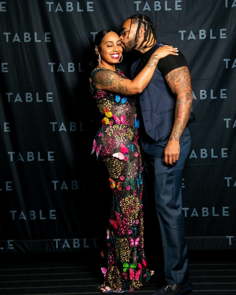 Couple posing for a photo at an event with a black backdrop with the word "TABLE" repeated.