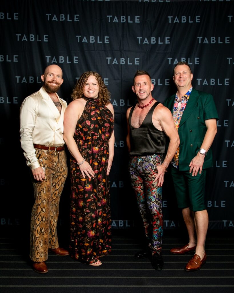 Four people pose in front of a "TABLE" backdrop at a formal event.