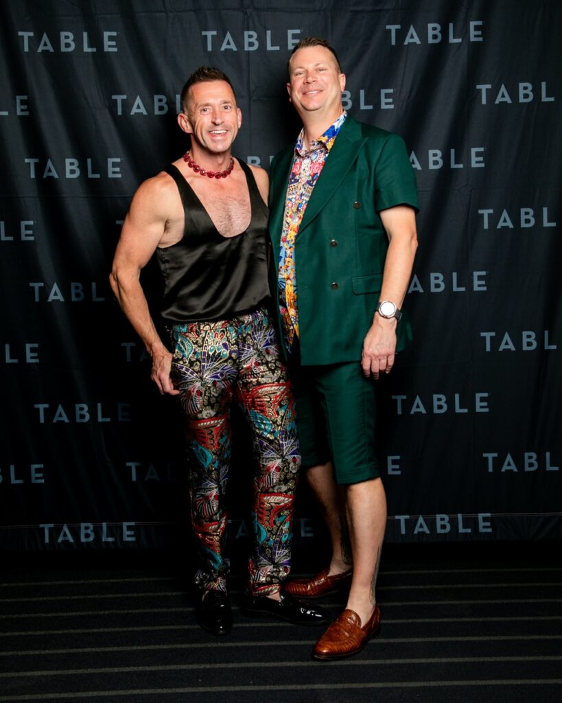 Two men pose in stylish outfits in front of a black backdrop with repeating "TABLE" text.