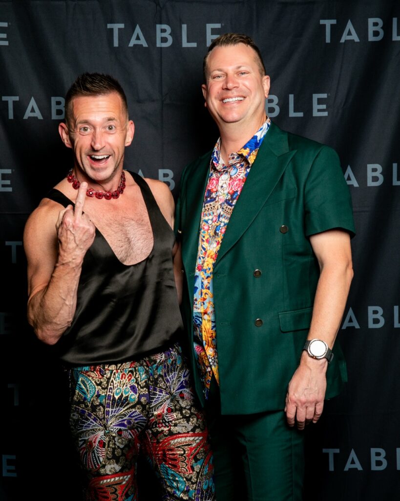 Two men in colorful outfits pose in front of a black backdrop with "TABLE" logo.
