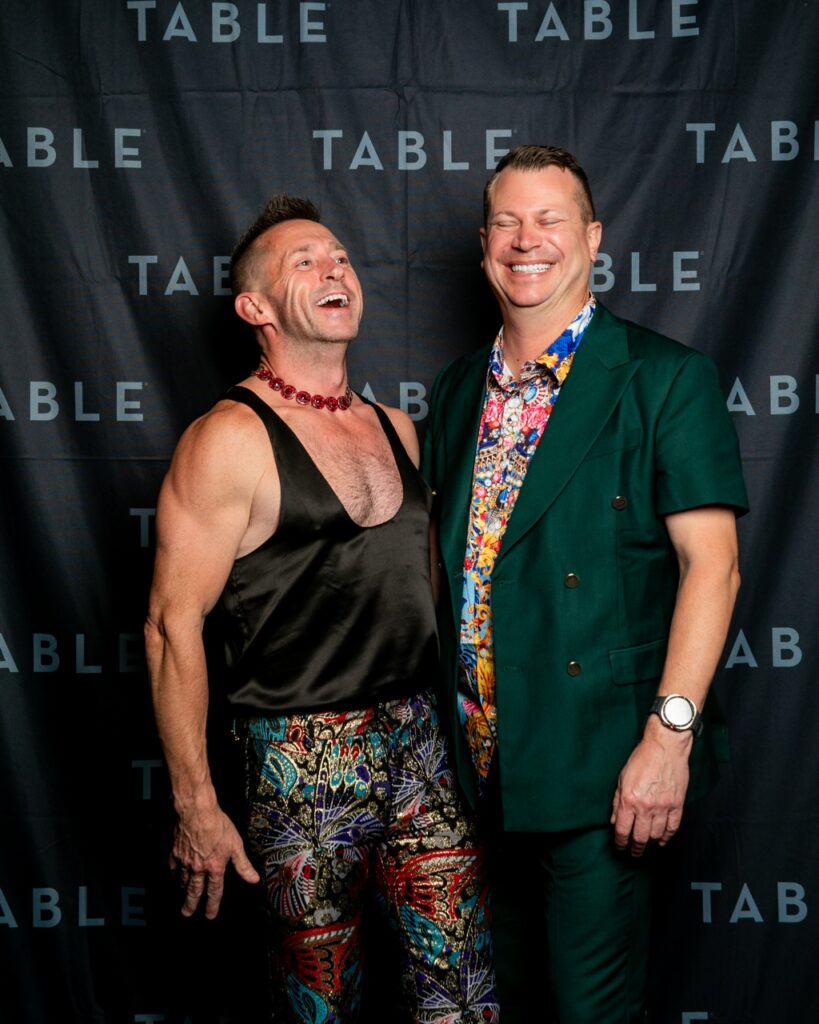 Two men in colorful outfits laugh in front of a black backdrop with "TABLE" repeatedly printed on it.