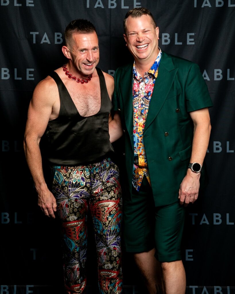 Two men in vibrant, fashionable outfits pose for a photo at an event with a "TABLE" backdrop.