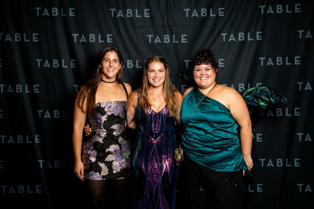 Three women smile in front of a black backdrop with "TABLE" repeatedly printed on it.