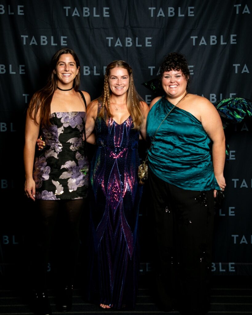 Three women in formal attire pose for a photo in front of a TABLE backdrop.