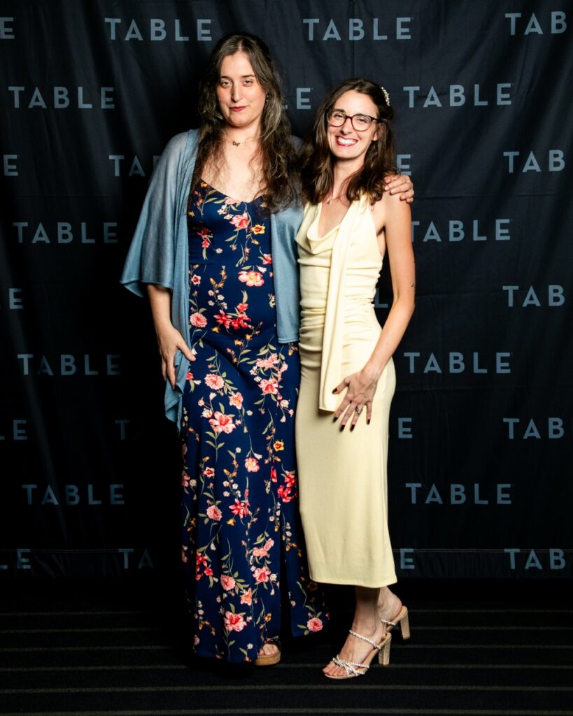 Two women smile for a photo in front of a TABLE backdrop.