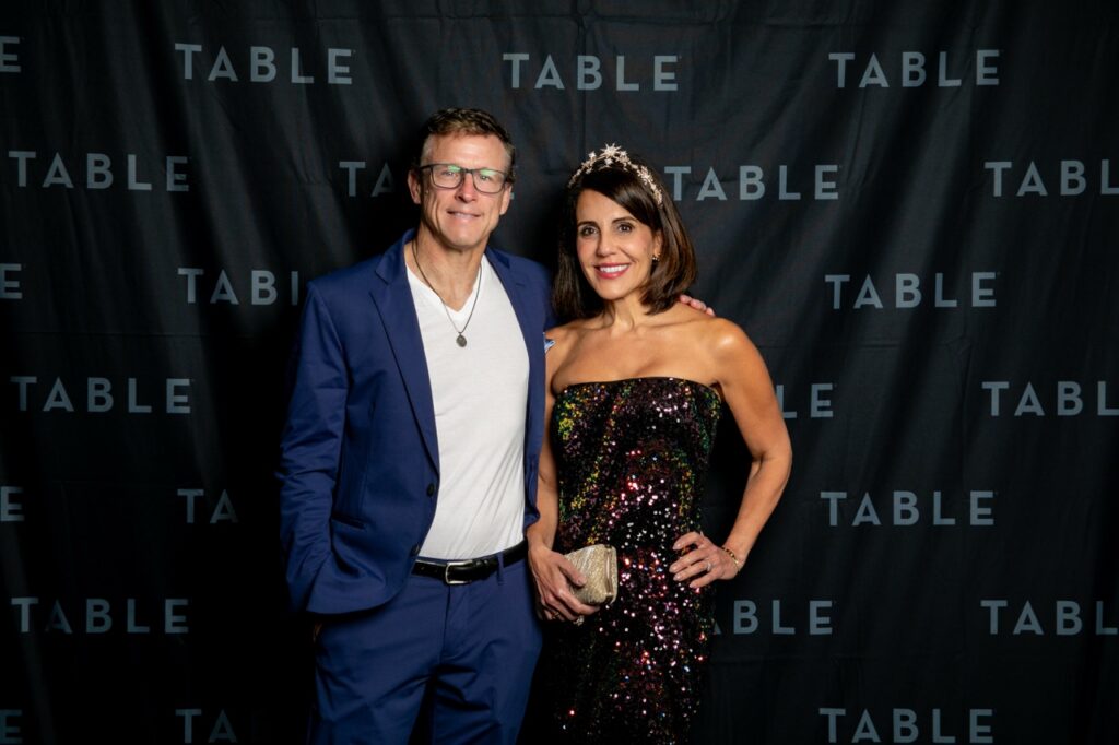 Man in blue suit and woman in sequin dress pose in front of a black TABLE backdrop.