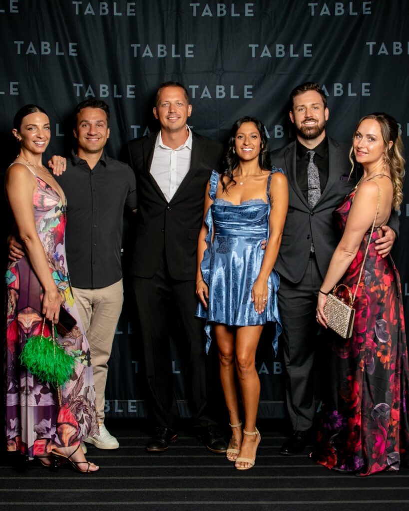 Six people pose for a photo in front of a black backdrop with the word "TABLE" repeated.
