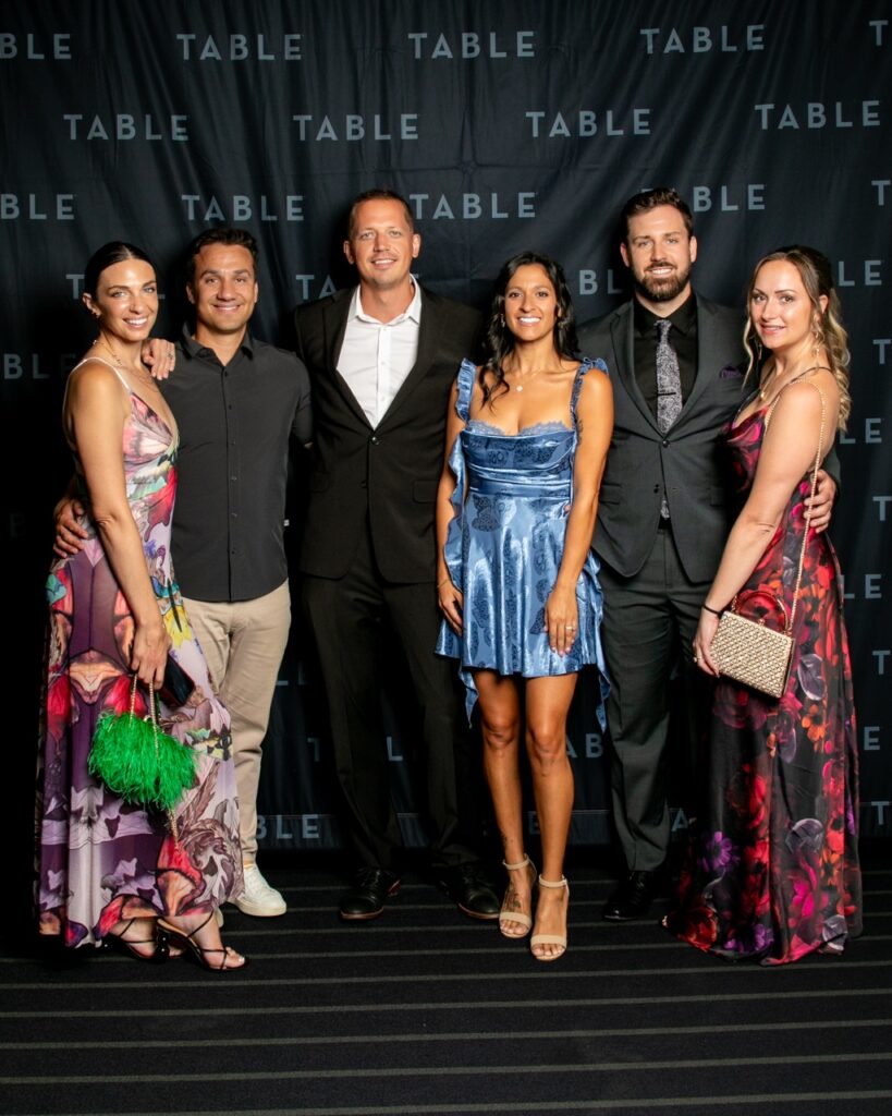 Six people in formal wear pose for a photo in front of a black backdrop with the word "TABLE" repeated.