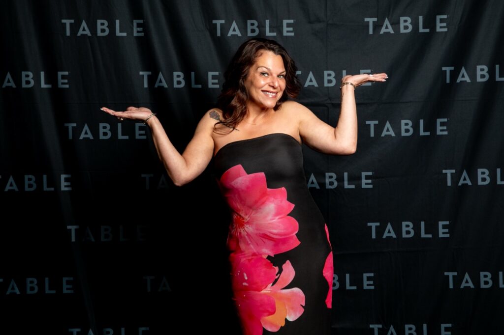 Woman in floral dress poses with arms outstretched against a black backdrop with "TABLE" logo
