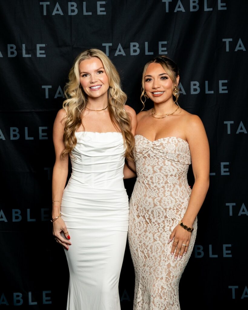 Two women in formal dresses pose in front of a black backdrop with the word "TABLE" repeated.