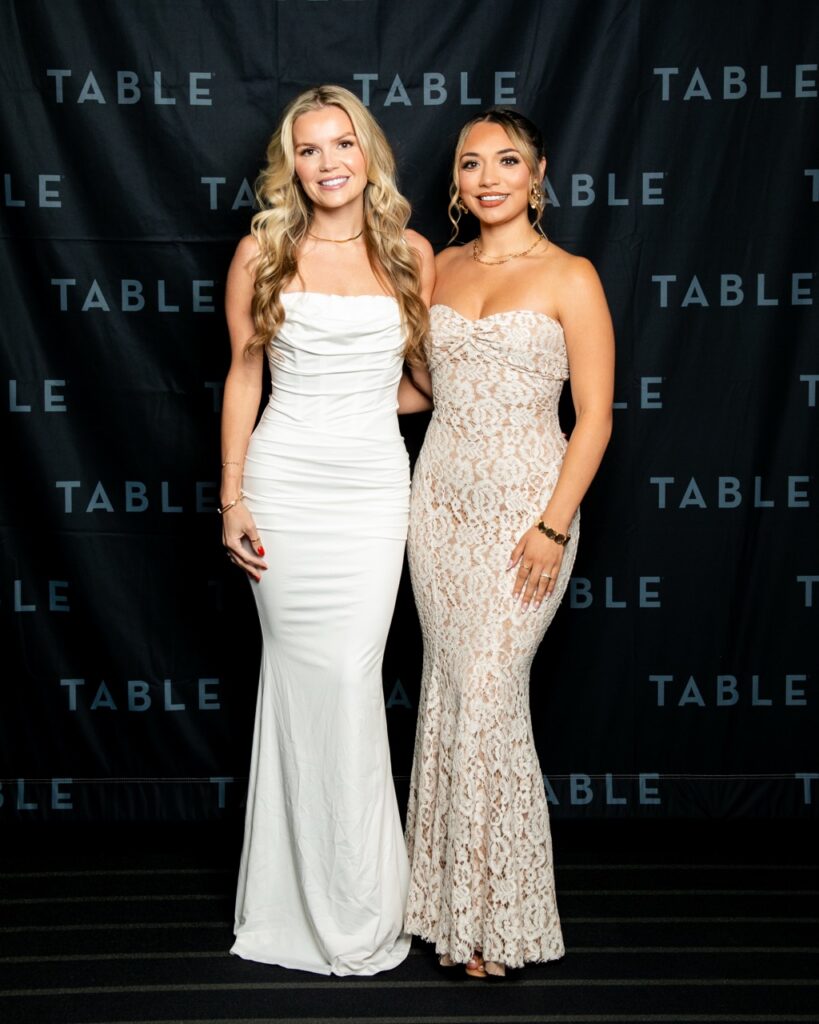 Two women in formal gowns pose in front of a backdrop with "TABLE" repeated.