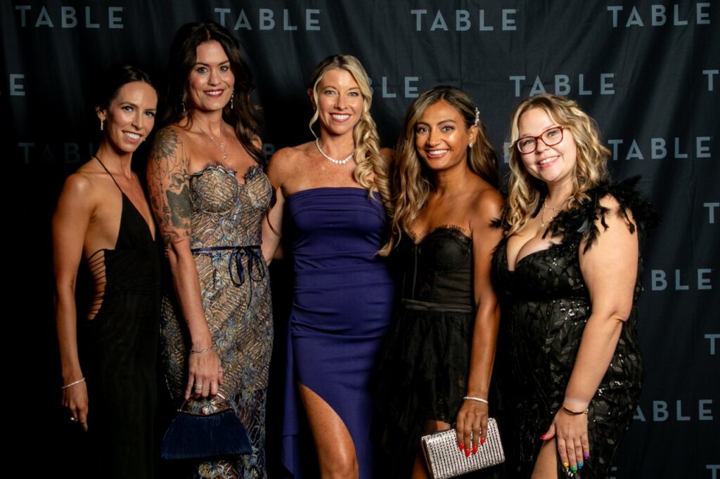 Five women in formal dresses pose for a photo at an event with a TABLE backdrop.