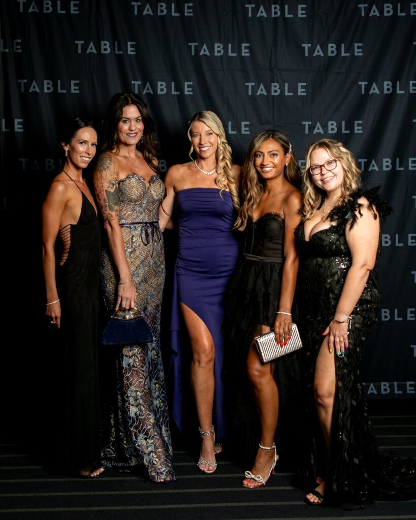 Five women in formal gowns pose for a photo at an event with a "TABLE" backdrop.