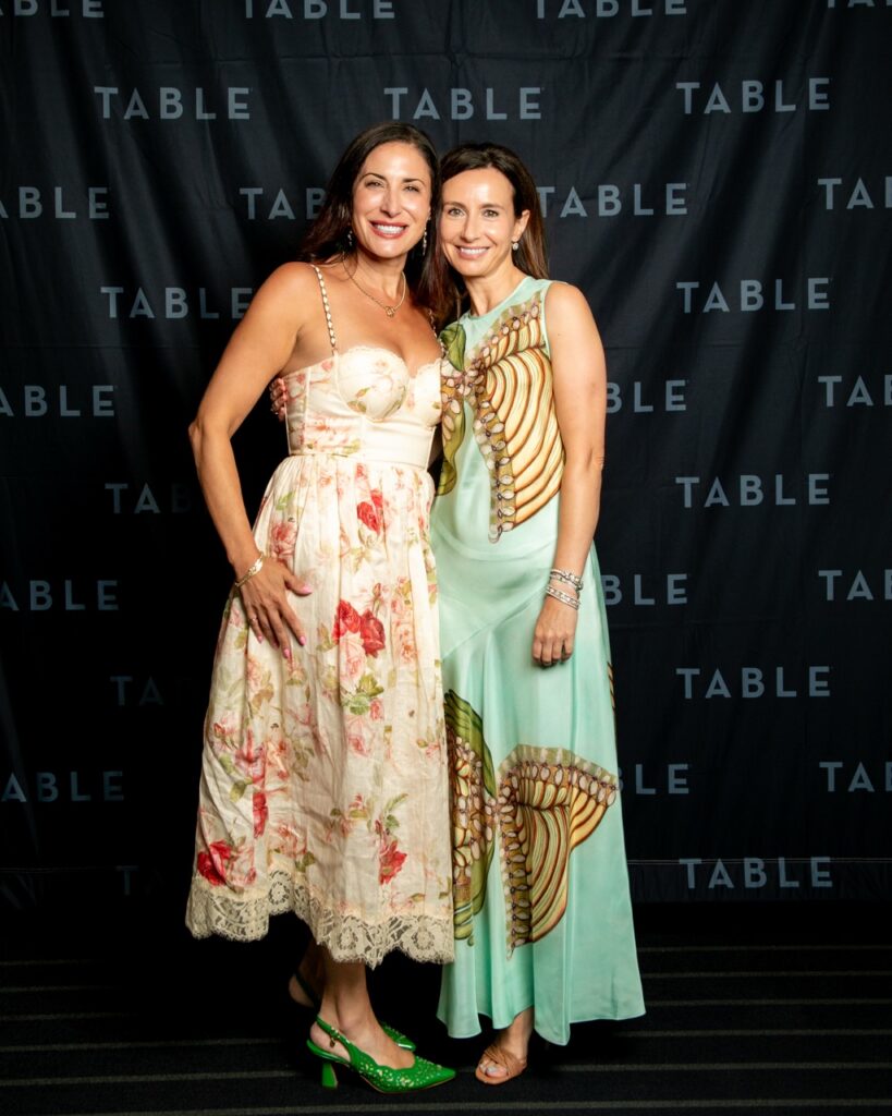 Two women in elegant dresses pose in front of a black backdrop with "TABLE" repeated.