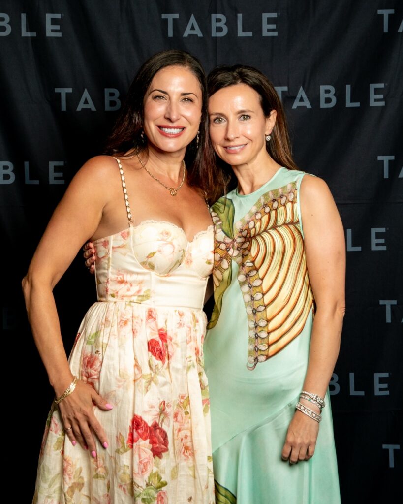 Two women pose for a photo in front of a black backdrop with the word "TABLE" repeated.