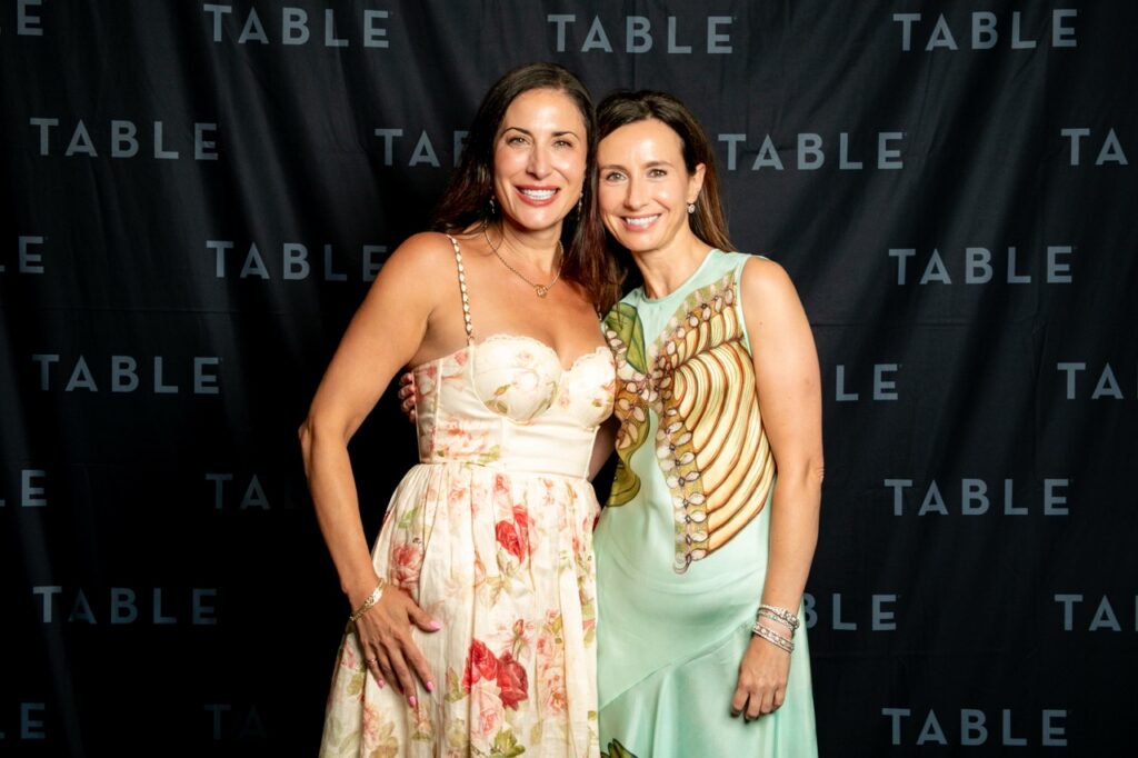 Two women in formal attire smile in front of a black backdrop with "TABLE" repeated.