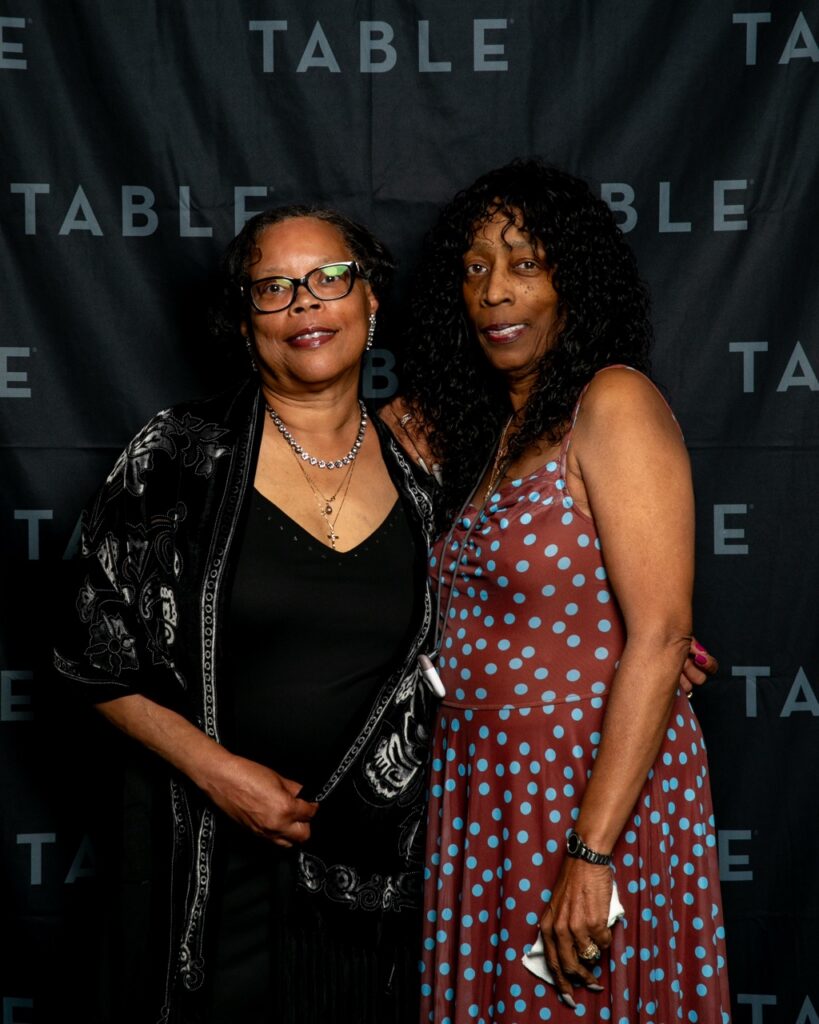 Two women posing for a photo in front of a black backdrop with the word "TABLE" repeated.