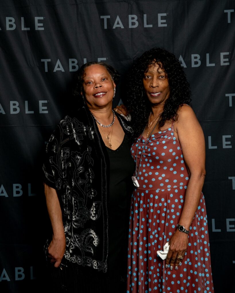 Two smiling Black women pose in front of a black backdrop with "TABLE" logo