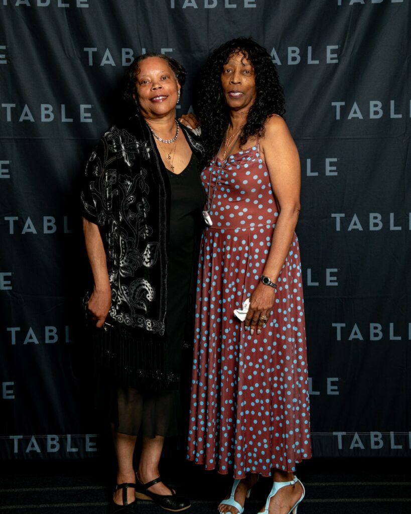 Two Black women pose for a photo in front of a black backdrop with repeating "TABLE" text.