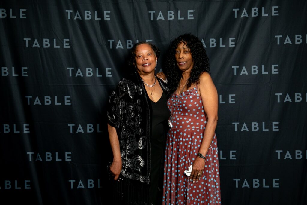 Two women in formal attire pose for a photo in front of a black backdrop with "TABLE" printed repeatedly.