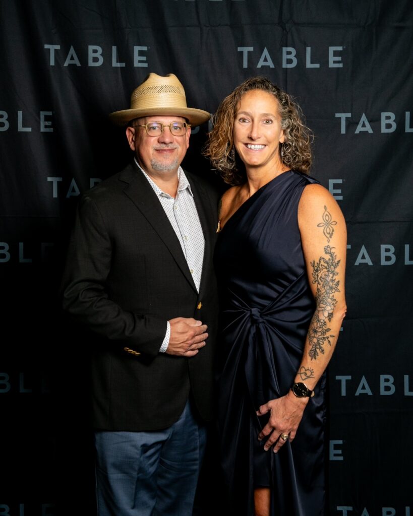 Man in hat and woman in navy dress pose in front of a TABLE backdrop