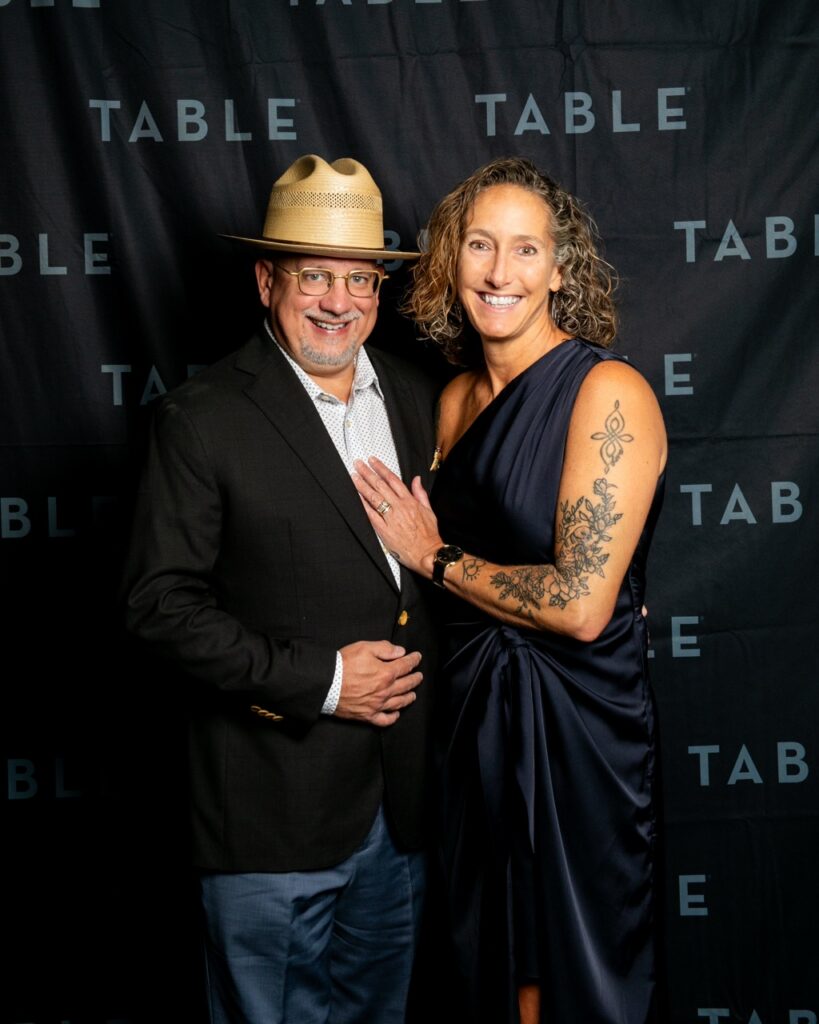 Man in fedora and woman in navy dress pose in front of "TABLE" backdrop.