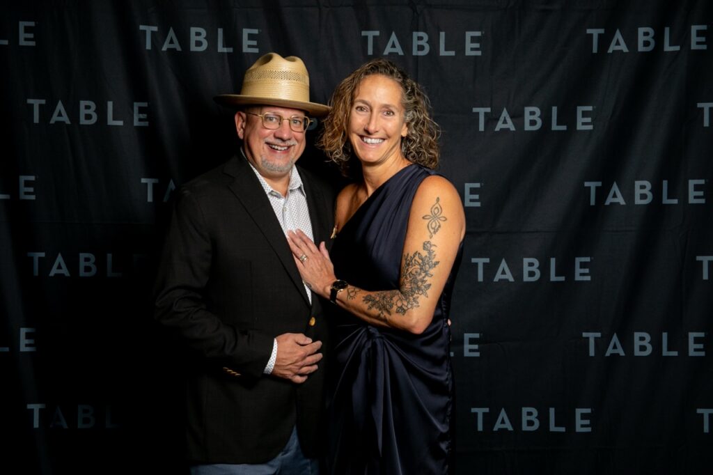 Couple posing at a TABLE event, man in hat, woman with tattoo