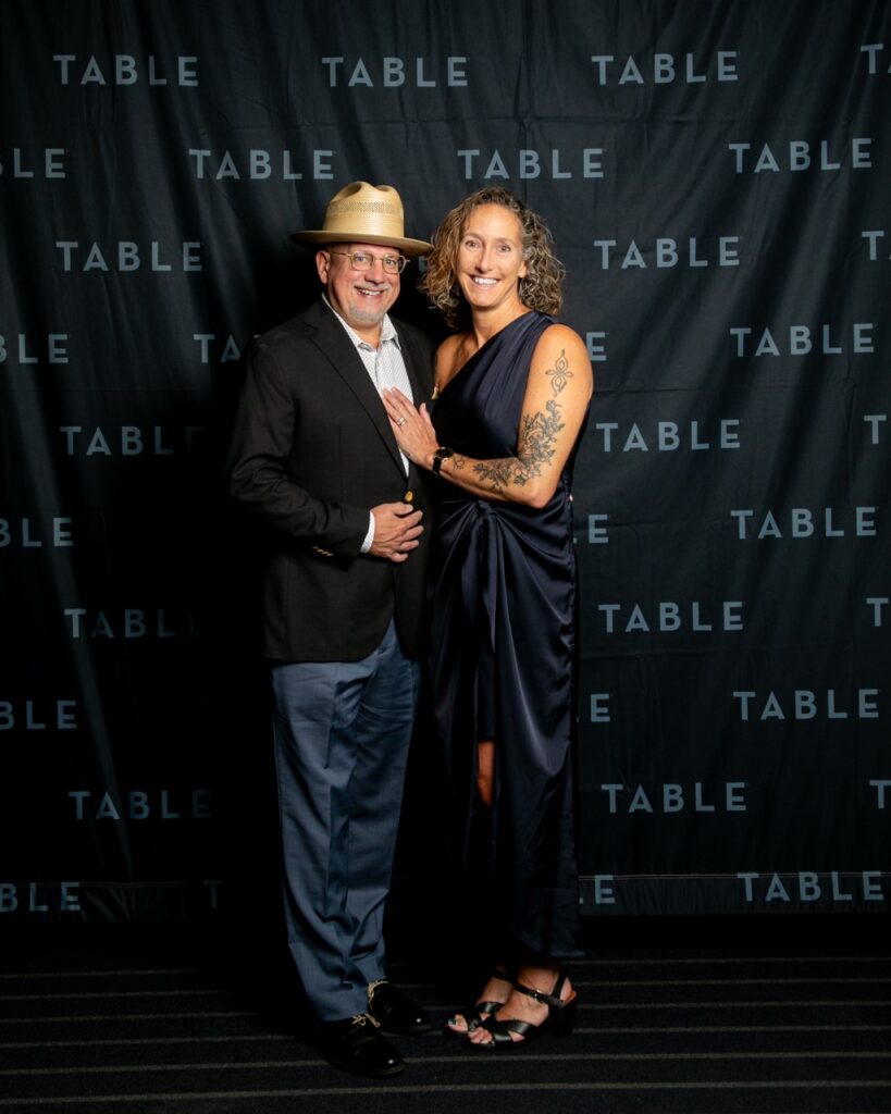 Couple posing in front of a black backdrop with repeating "TABLE" logo.