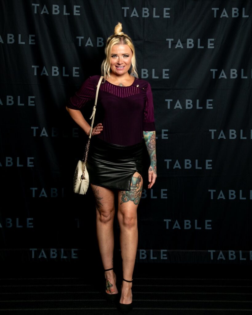 Woman in purple top and leather skirt with tattoos poses for a photo.