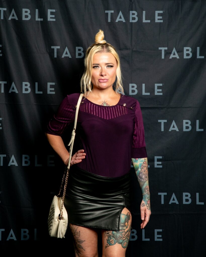 Woman with tattoos in a purple top and black skirt poses in front of a "TABLE" backdrop.