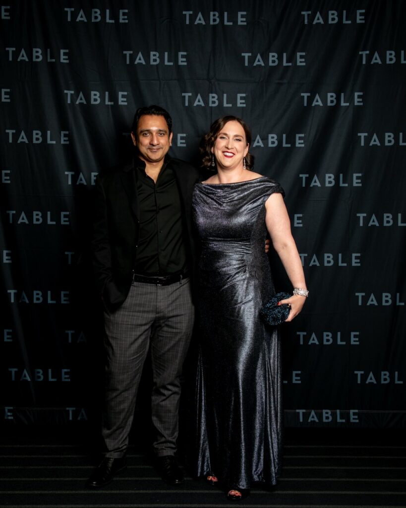 Couple posing in front of a black backdrop with repeated "TABLE" text.