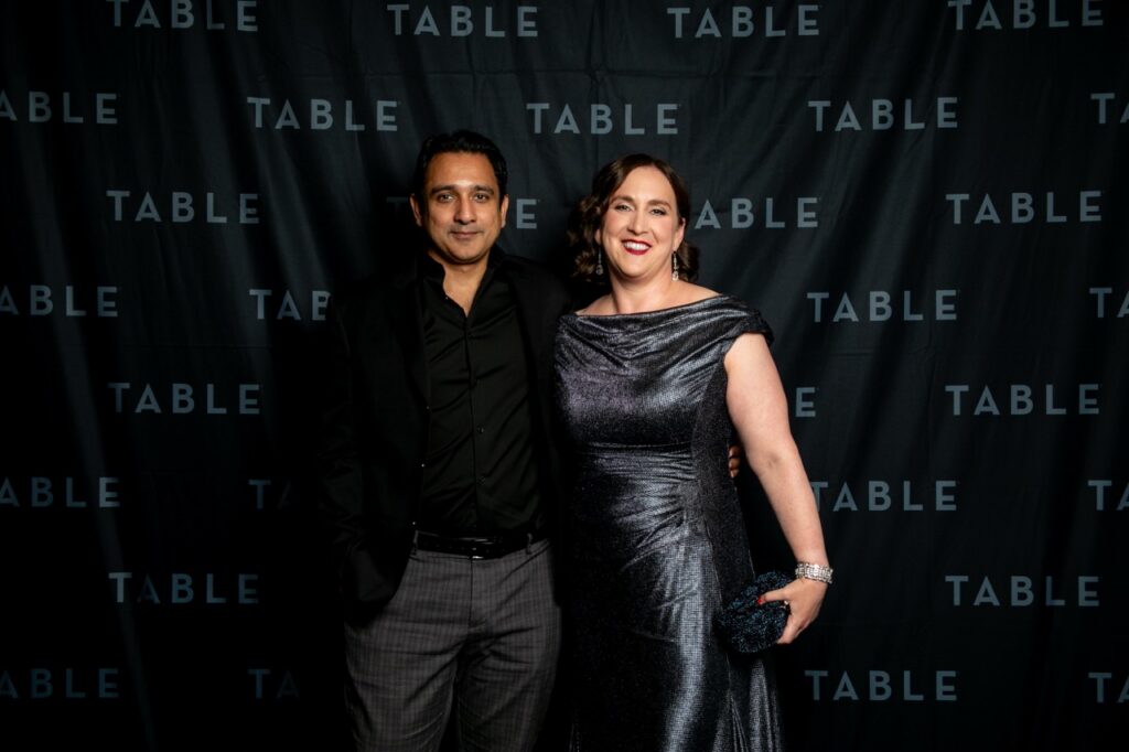 Couple posing in front of a black backdrop with "TABLE" repeated