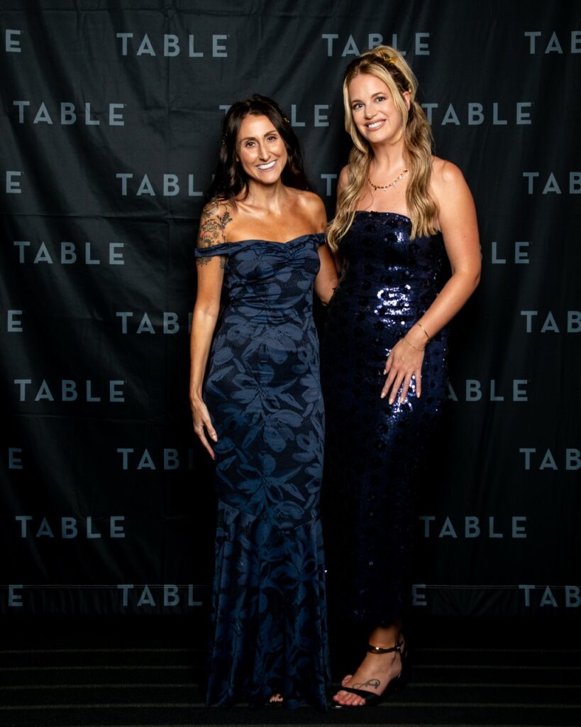 Two women in elegant navy dresses pose in front of a black backdrop with the word "TABLE" repeated.
