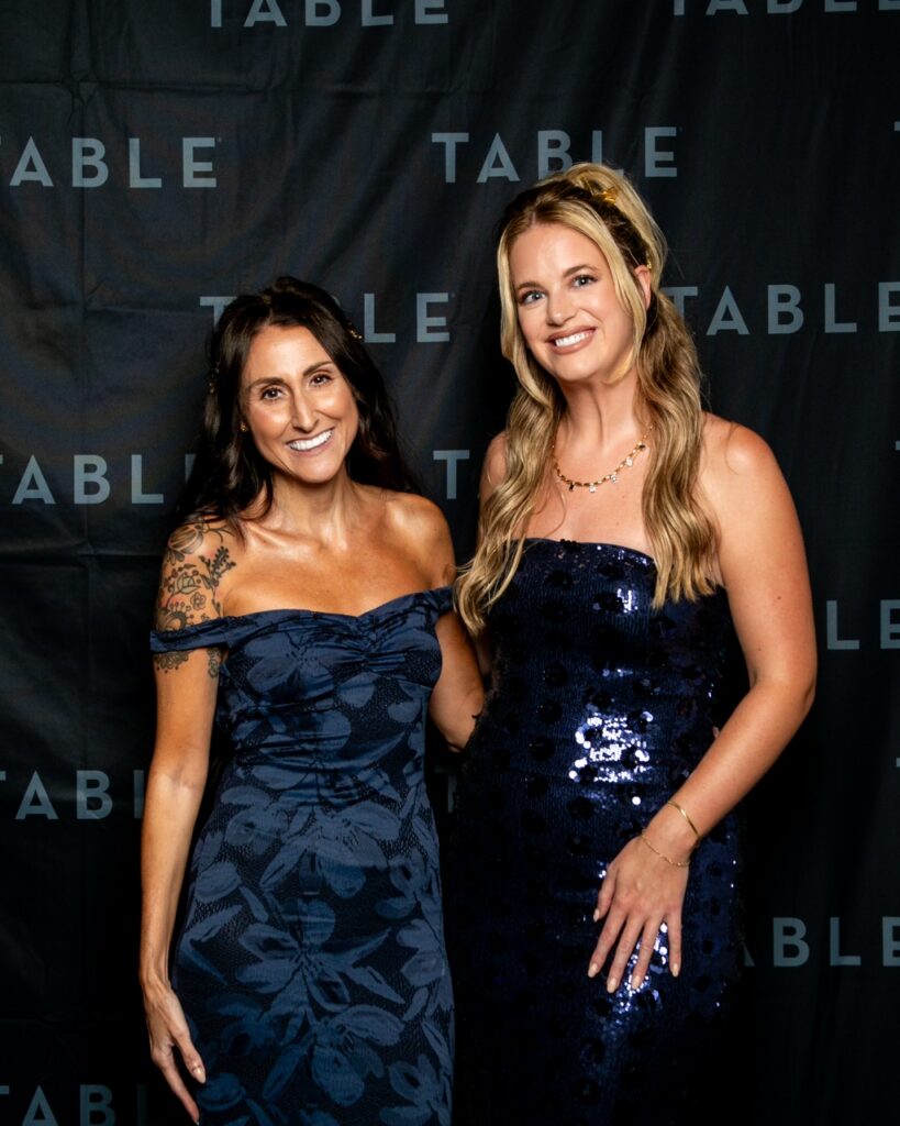 Two women in navy blue dresses pose for a photo in front of a black backdrop with the word "TABLE" repeated.